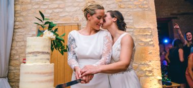 Two Lesbian Brides Cutting Cake And Kissing At Upcote Barns In Cotswolds Via Gay Wedding Guide By Noel Hibbert 9 380X174