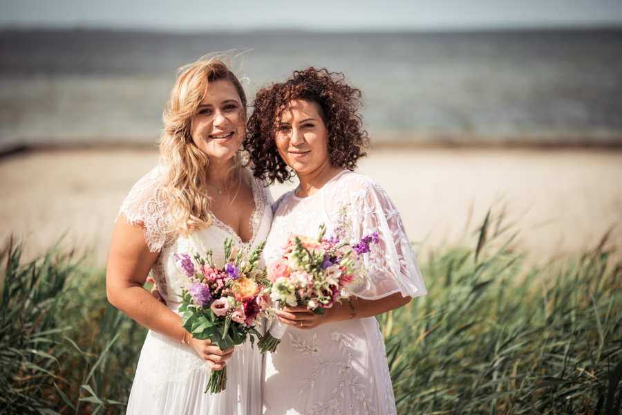 A couple smiling and holding bouquets a Nordic beach during their gay wedding in Denmark.